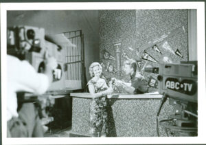 Photograph, showing ABC television studios, paper, photographed by Bob Eaton, printed by Kodak, Sydney, New South Wales, Australia, 1959-1961 Black and white landscape-format photographic print with white border. The photograph features Johnny O'Keefe conversing with a woman on set at ABC's Gore Hill television studios. The text 'Six O'Clock Rock' is handwritten in lead pencil on the reverse side of the image.